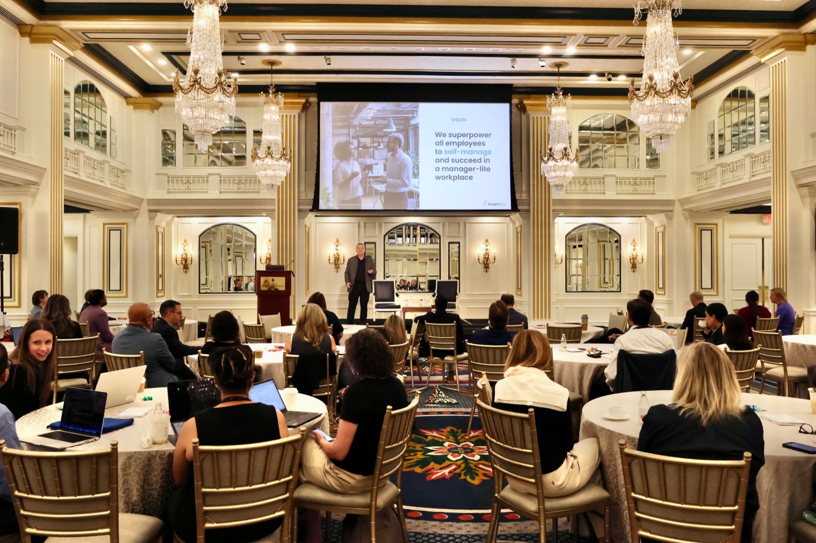 Alberto delivering a Tough Day keynote in a gilded ballroom with crystal chandeliers, presenting a slide titled 'We superpower all employees to self-manage and succeed in a manager-lite workplace'